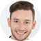 A young man with short brown hair and trimmed beard smiles broadly at the camera against a white background.