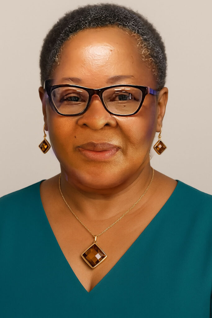 A woman with short gray hair wearing glasses, a teal top, and matching gold jewelry with square brown gemstones—earrings and a necklace—smiling softly against a neutral background.